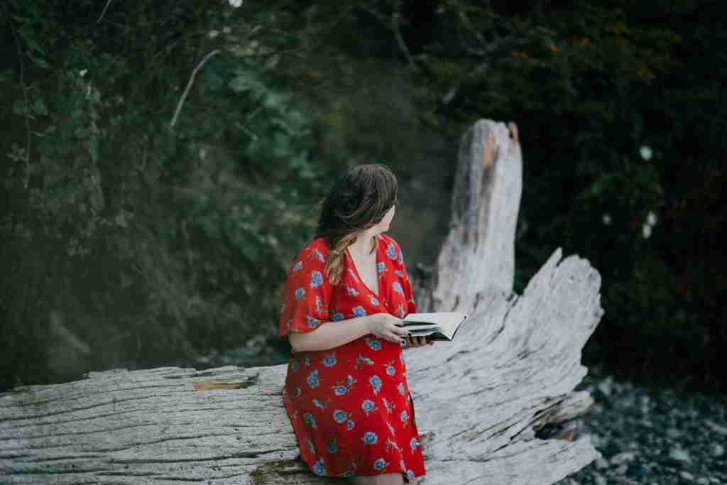 Una mujer sentada en un tronco caído en un bosque, con un vestido rojo de flores, sosteniendo un libro abierto y mirando a un lado. La escena, serena y reflexiva, refleja el vibrante color de la Iglesia para el Domingo de Ramos.