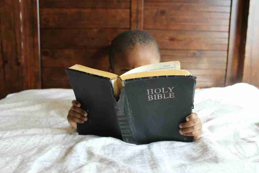 Uma criança está sentada em uma cama com cabeceira de madeira, profundamente absorta na leitura da Bíblia Sagrada, talvez se preparando para a aula da Escola Dominical sobre a Armadura de Deus.
