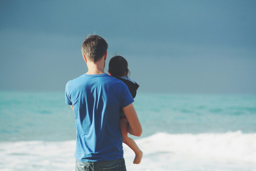 A man in a blue shirt holds a child, like a guardian from stories of orphans in the Bible, facing the vast ocean with waves crashing gently in the background.