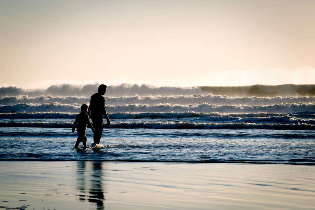 Silhouetted adult and child walking along the beach, reminiscent of stories of orphans in the Bible, with waves in the background under a bright sky.
