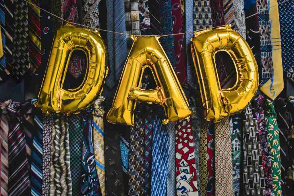 Letras douradas em forma de balão com a inscrição "DAD" estão penduradas sobre uma variedade colorida de gravatas, perfeitas para ideias de presentes para o Dia dos Pais.