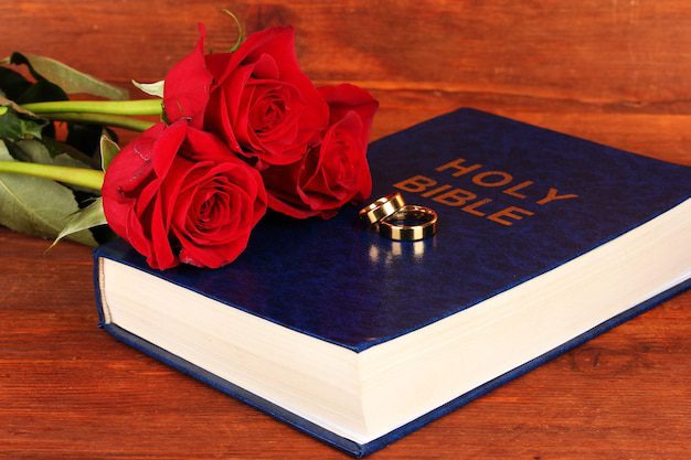 Una Biblia con anillos dorados y rosas rojas descansa sobre una superficie de madera, simbolizando la unión duradera del hombre y la mujer en las enseñanzas intemporales de la Biblia.