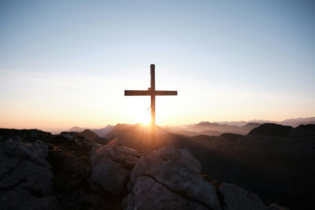 A silhueta de uma cruz no pico de uma montanha é iluminada pelo sol que se põe atrás de montanhas distantes, evocando um momento de serena consagração, conforme descrito na Bíblia.