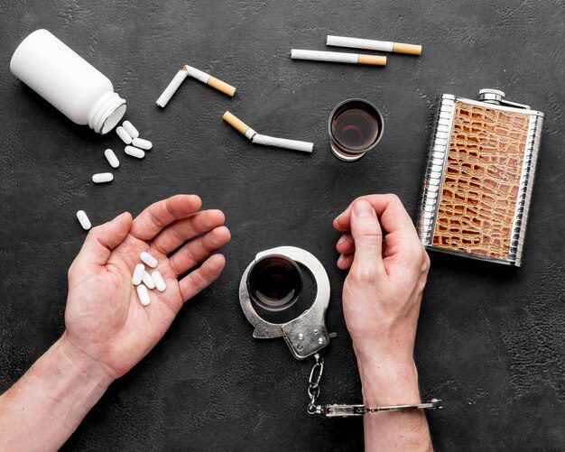 Um indivíduo algemado agarra comprimidos, cercado por cigarros, álcool e algemas em uma superfície sombria, evocando a contemplação das correntes da vida. É uma cena dura que convida à reflexão sobre o controle da dependência - um momento que nos leva a perguntar "O que a Bíblia diz sobre a dependência de drogas?