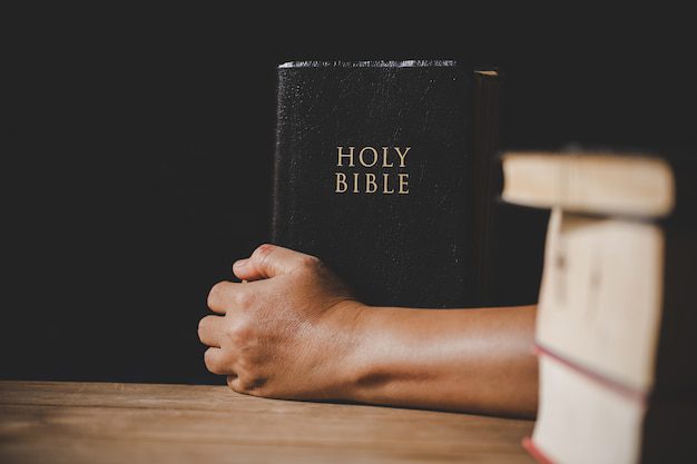Um indivíduo segura uma Bíblia Sagrada preta com uma superfície de madeira, ponderando sobre a antiga questão: De onde veio a esposa de Caim na Bíblia? Livros borrados estão em primeiro plano, acrescentando um ar de mistério a essa investigação bíblica.
