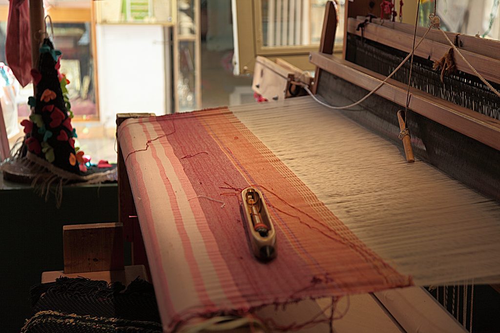 Em uma sala iluminada pelo sol, um tear manual tece tecidos vermelhos e brancos com uma lançadeira de madeira, ecoando o artesanato dos tempos bíblicos - talvez uma reminiscência das histórias antigas como a de Boaz, que se casou com Rute na Bíblia.