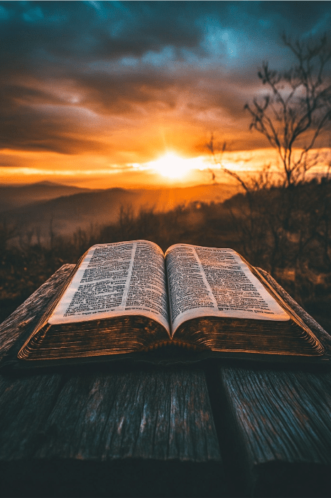 Um livro aberto repousa sobre uma mesa de madeira, enquanto o pôr do sol lança um brilho quente sobre montanhas distantes, convidando à reflexão e evocando a contemplação sobre o que a Bíblia diz sobre o destino.