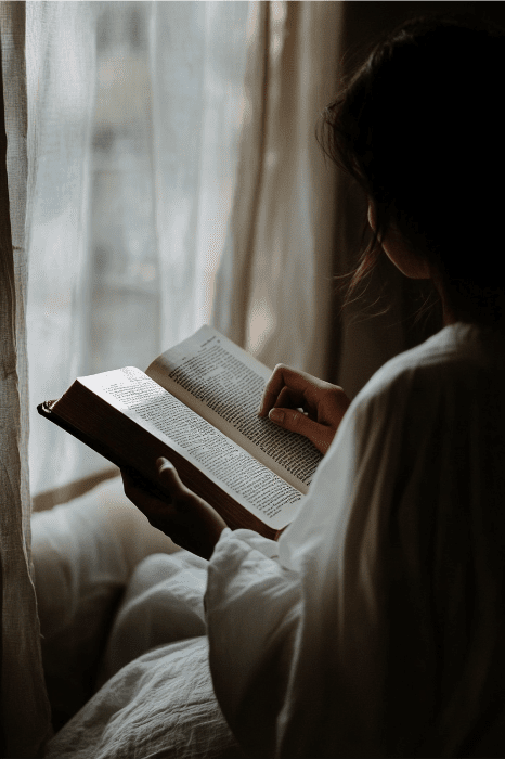 A person in a white shirt reads a book by a window with sheer curtains, creating a soft, natural light atmosphere. It's a tranquil moment that prompts reflection on the question: What does the Bible say about reading the Bible?.