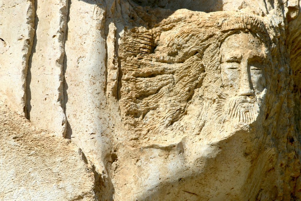 An ancient stone carving of a bearded man's face, reminiscent of biblical figures whose names prompt the question: "I Am in the Bible How Many Times?" etched into a rough rock surface.