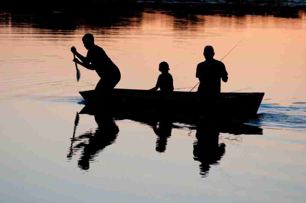 Silhouettes of three people in a boat, reminiscent of the Fisher of Men parable, enjoy fishing at sunset on a calm lake.