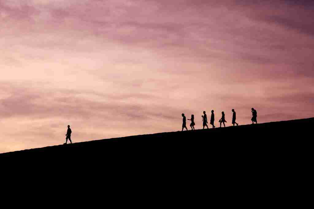 Silhuetas de pessoas, que lembram Moisés guiando seus filhos, caminham em fila em uma colina contra um céu rosa e roxo ao pôr do sol.