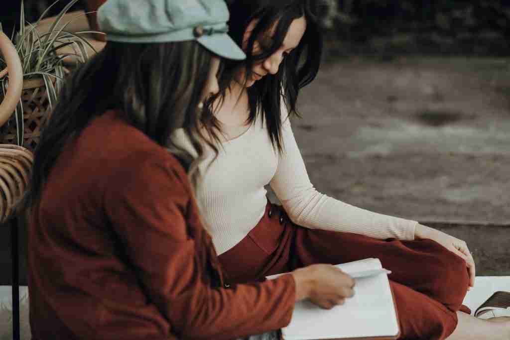 Two people sit outdoors, one engrossed in a book. The person on the left wears a cap, reminiscent of a modern-day fisher of men, while the other curiously looks at the Bible's pages.