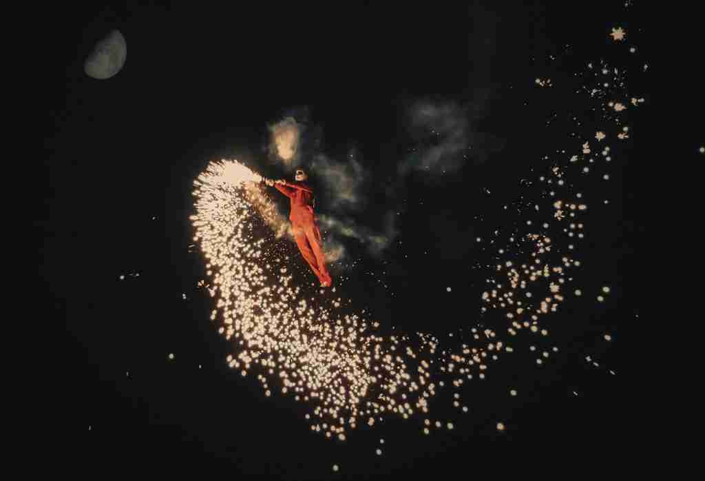 Uma pessoa em um terno vermelho realiza uma deslumbrante exibição de acrobacias com fogos de artifício à noite, cercada por faíscas em forma de meia-lua, lembrando o fascínio hipnotizante do Espírito de Jezabel na Bíblia.