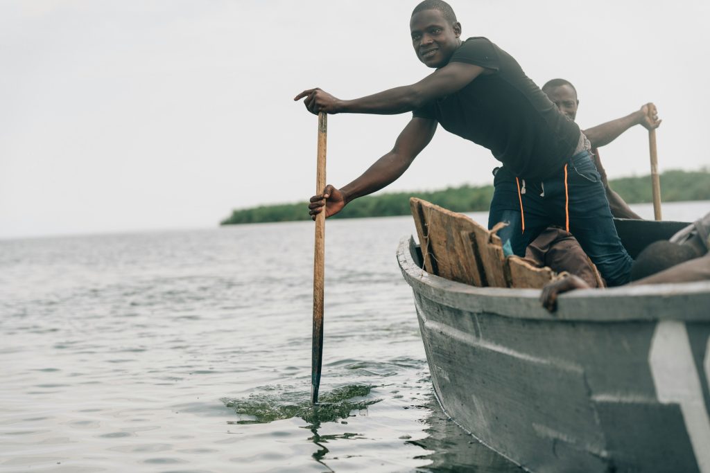 Dos hombres, como los Hijos del Trueno de la Biblia, reman una barca de madera en una tranquila masa de agua, con la vegetación formando el sereno telón de fondo.