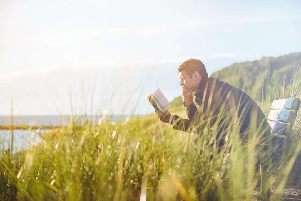 Man reading gentleness Bible verses on a bench surrounded by tall grass with a scenic view in the background.