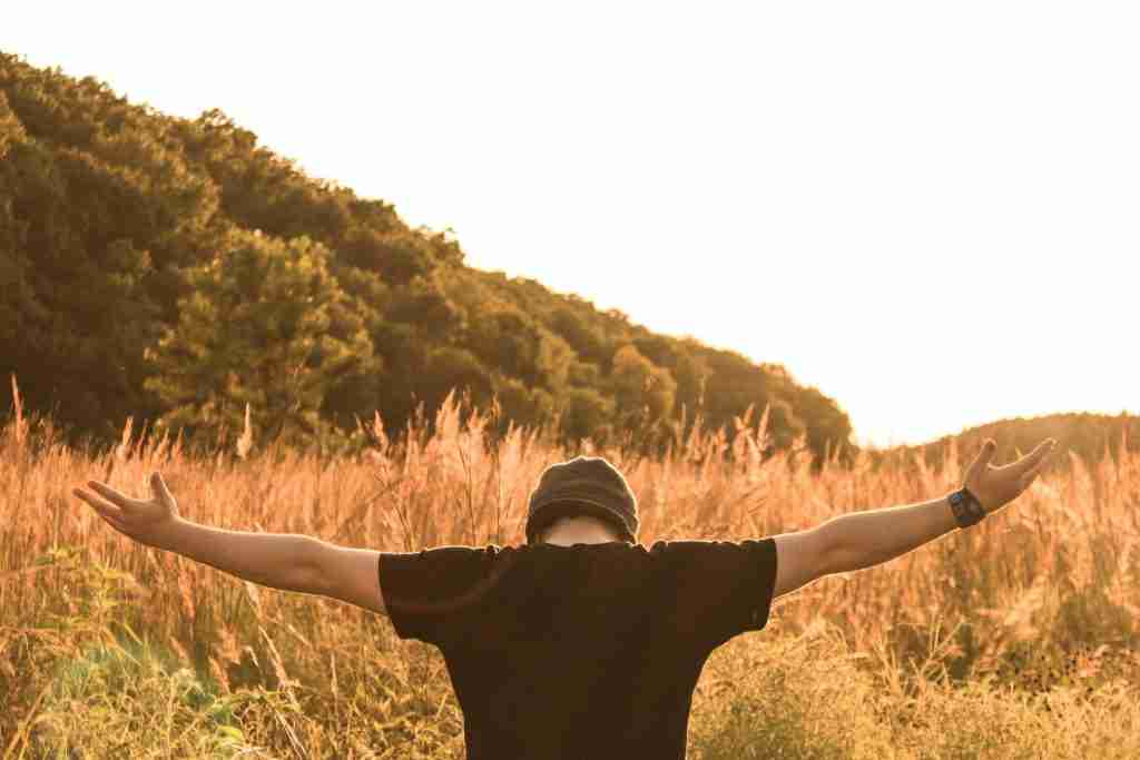 Persona en un campo con los brazos extendidos, frente a una brillante puesta de sol con colinas y árboles al fondo, reflejando la serenidad que se encuentra en las oraciones de Alcohólicos Anónimos.
