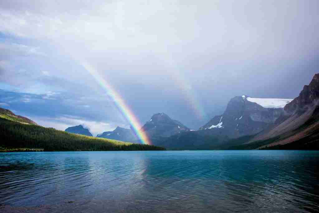 Doble arco iris sobre un sereno lago de montaña con picos nevados al fondo y una costa boscosa, invocando la paz que se encuentra en las Oraciones de Alcohólicos Anónimos.
