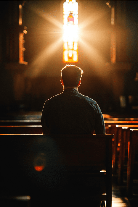 Silhouetted person sitting on a pew in a dimly lit church, lost in deep prayers, as sunlight streams through a stained glass window.