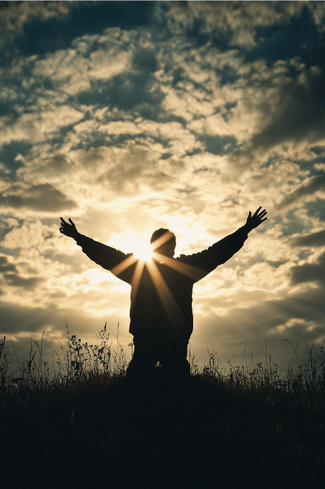 Pessoa em silhueta com os braços erguidos contra o sol poente e o céu nublado em uma colina gramada, oferecendo mensagens curtas de reflexão para reuniões de oração.