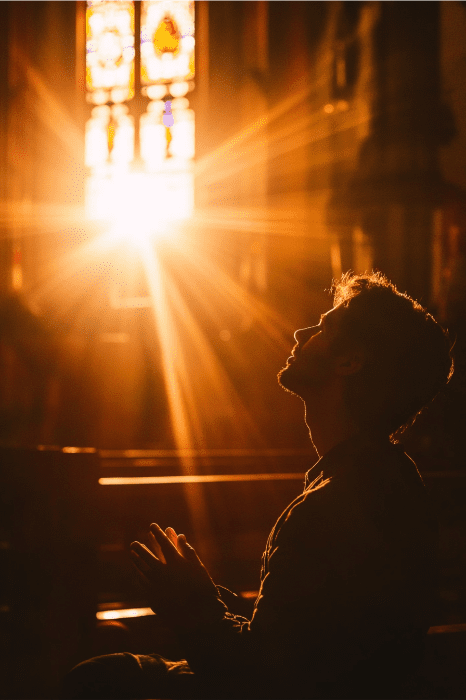In the dimly lit room, sunlight streams through a stained glass window, casting vibrant hues on the silhouette of a person deep in prayer for revival in the church.
