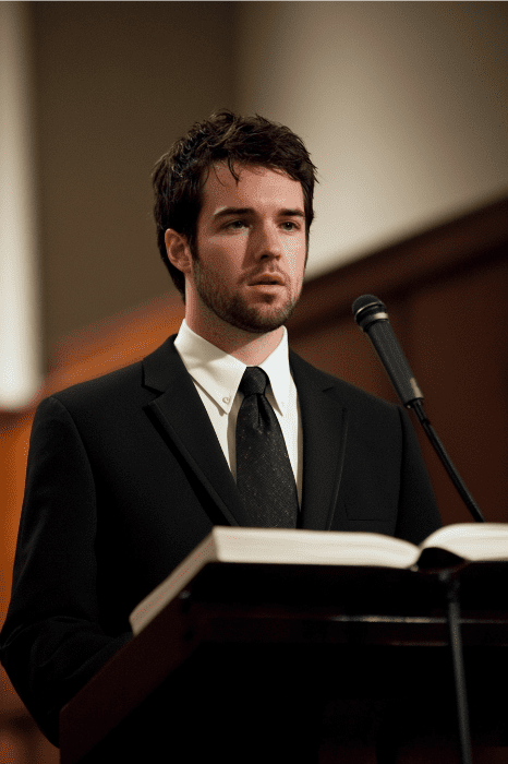 A man in a suit delivers an expository sermon series, speaking passionately at a podium with an open book, microphone in hand, captivating his audience in the formal setting.