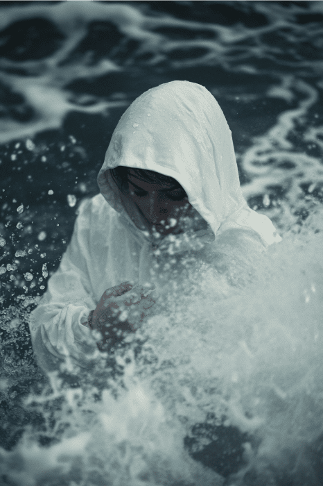Em meio a respingos de água, uma pessoa com um capuz branco está de pé, parcialmente obscurecida pelas ondas, evocando ideias ponderadas da série de sermões sobre como navegar pelas marés turbulentas da vida.