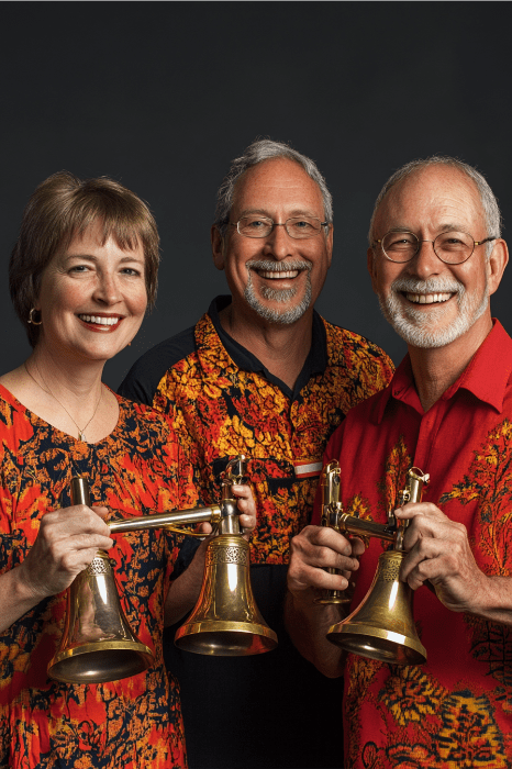 Three people smiling and holding handbells, wearing colorful patterned shirts against a dark background, showcasing one of the many joyful church volunteer ideas.