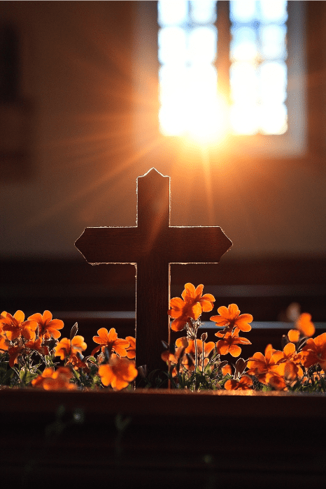 Silhueta de uma cruz cercada por flores alaranjadas, com a luz do sol passando por uma janela - um cenário sereno para ideias de sermões de Páscoa.