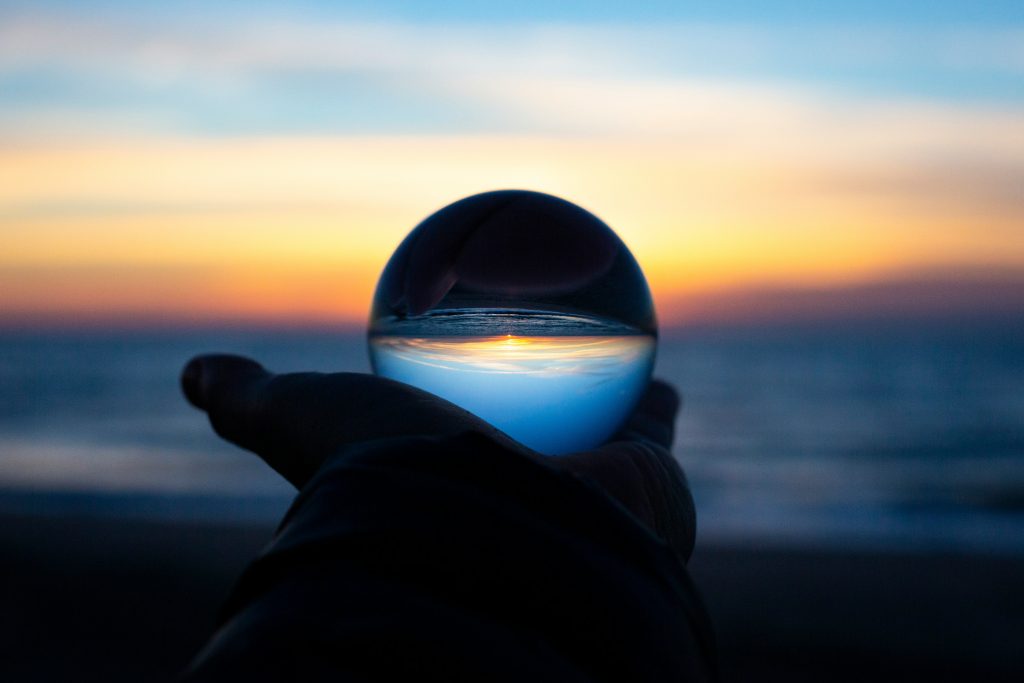 Uma bola de cristal erguida contra um pôr do sol sobre o oceano reflete o céu e o horizonte vibrantes, como se estivesse ponderando sobre o que uma profecia da Bíblia poderia revelar em um momento de tirar o fôlego.
