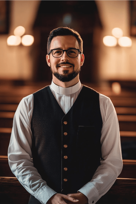 In a dimly lit church, a man wearing glasses, a clerical collar, and a vest stands smiling at the camera, perhaps contemplating valid reasons for changing churches.