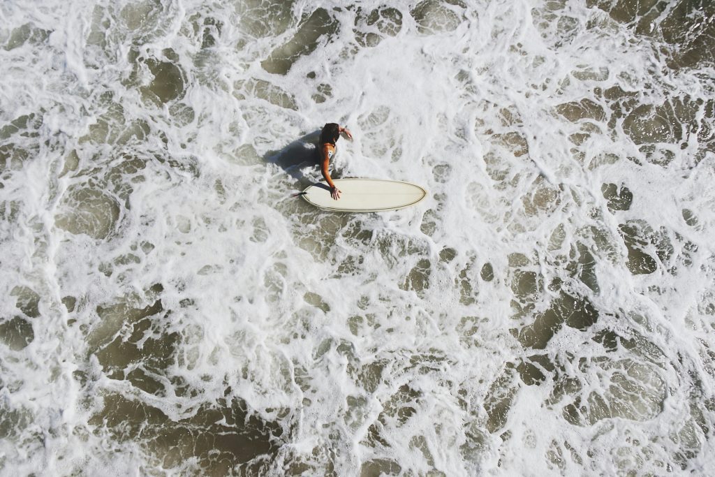 Viewed from above, a surfer with a white surfboard gracefully navigates through the foamy ocean waves, their movement akin to the perfect timing celebrated in the Bible.