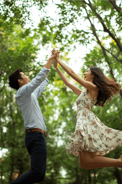 A joyful couple jumps and holds hands in a forest, surrounded by lush green trees, their laughter echoing Rebekah's spirit of adventure and joy as found in her story from the Bible.