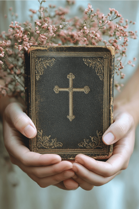 Hands holding an ornate book with a gold cross design, adorned with delicate pink flowers, perfect for reflecting on bible verses for sympathy cards.
