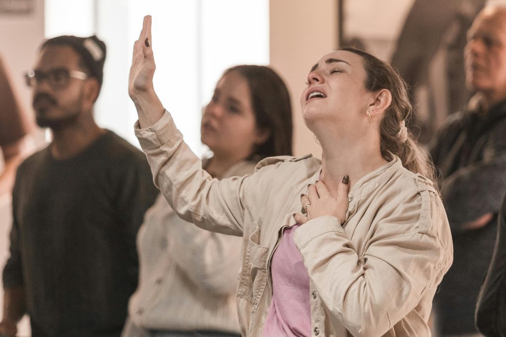 In the bustling crowd, a woman raises her hand, singing passionately with one hand on her chest, embodying the fervor of Nicodemus seeking truth in the night.