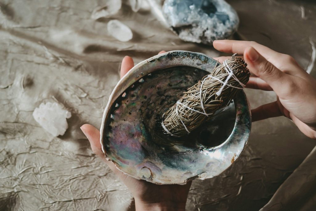 As mãos seguram uma concha de abalone com um feixe de sálvia, ecoando rituais como a queima de sálvia na Bíblia, cercada por cristais em uma superfície texturizada.