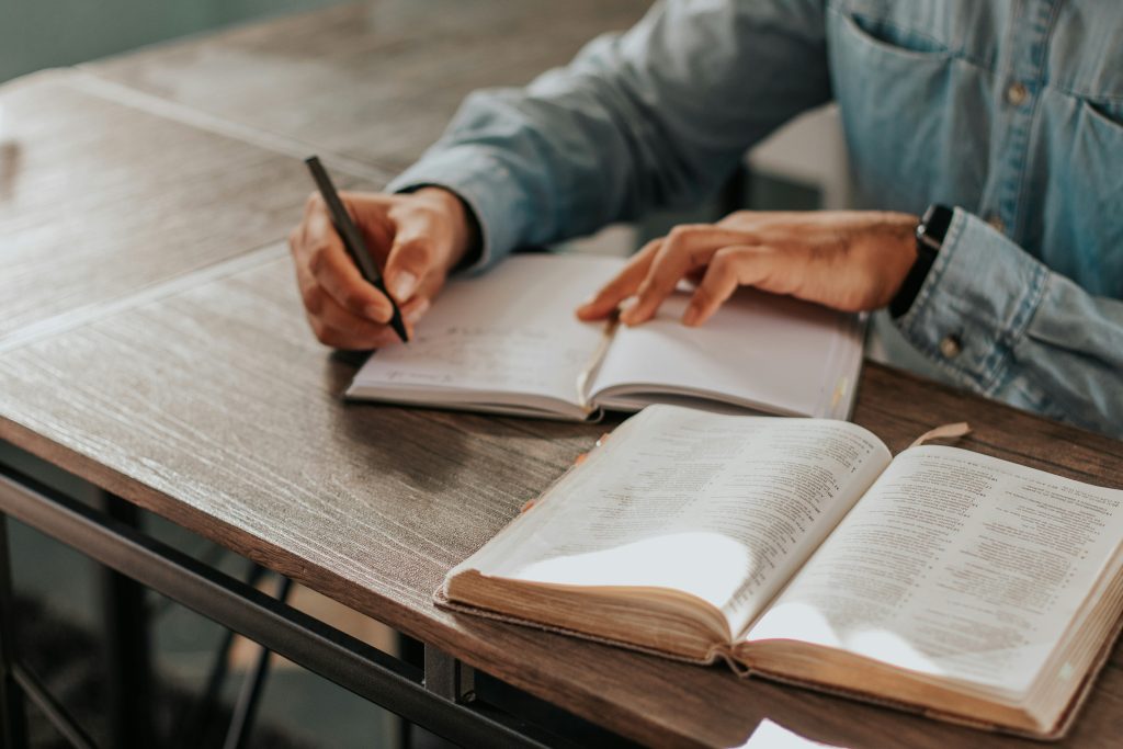 A person writing in a notebook with the Bible open next to them, studying perhaps pride in the Bible