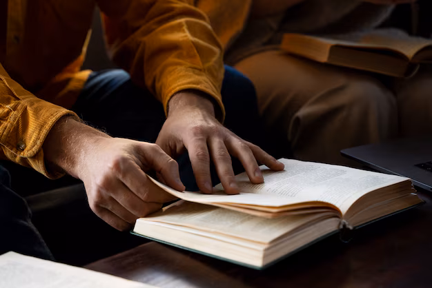 A person in a yellow shirt turns the page of an open book while reading at a table, perhaps pondering how many pages are in the Bible.