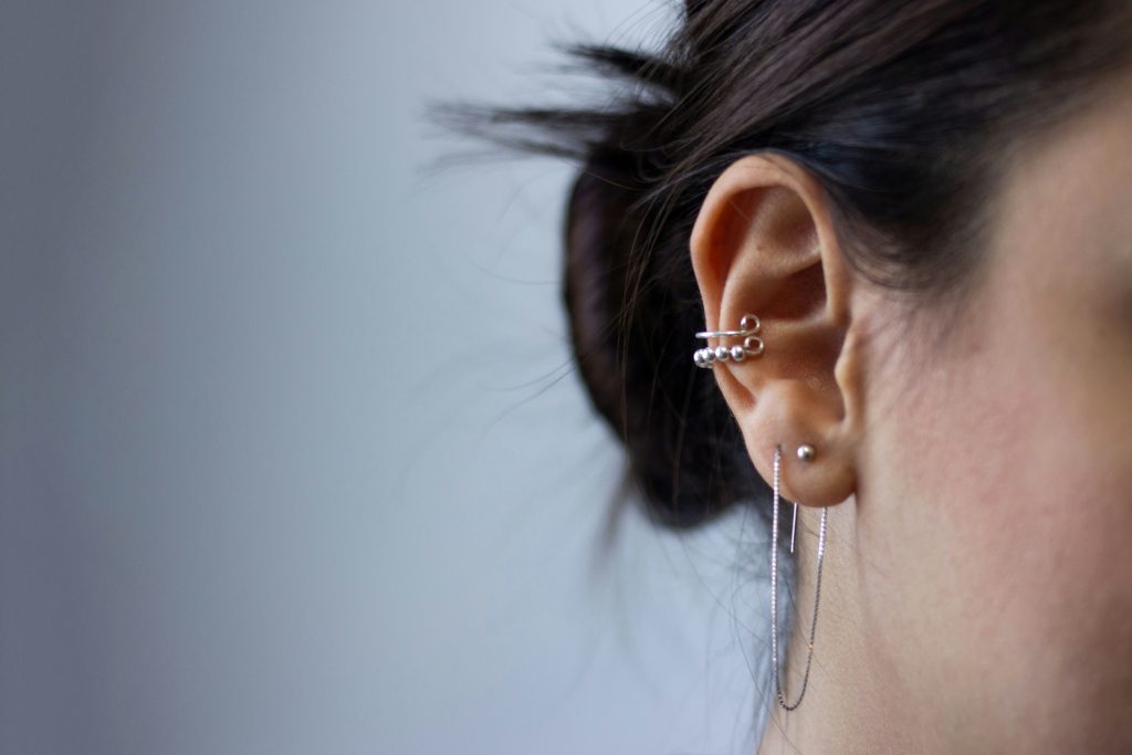 Close-up da orelha de uma pessoa com vários piercings, usando brincos de prata contra um fundo neutro, evocando uma sensação de ressonância espiritual semelhante ao zumbido místico nos ouvidos mencionado na Bíblia.