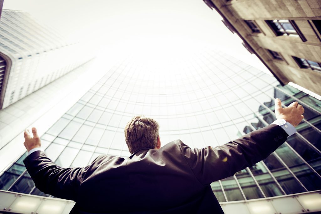 A man in a suit stands with arms stretched up towards skyscrapers, a sign of pride in the material things and not the Bible and God