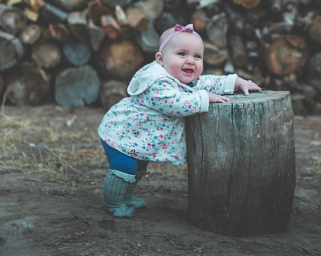 A baby in floral attire and blue leggings stands and smiles, leaning on a tree stump with logs in the background.