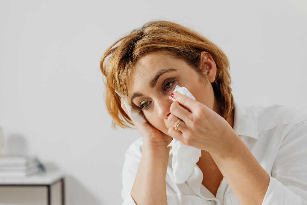 A woman, appearing upset or thoughtful, wipes her eye with a tissue. Dressed in a white shirt, she pauses in a light, minimalistic room. Thoughts linger like echoes of "What Happened to Hagar's Son Ishmael in the Bible?.