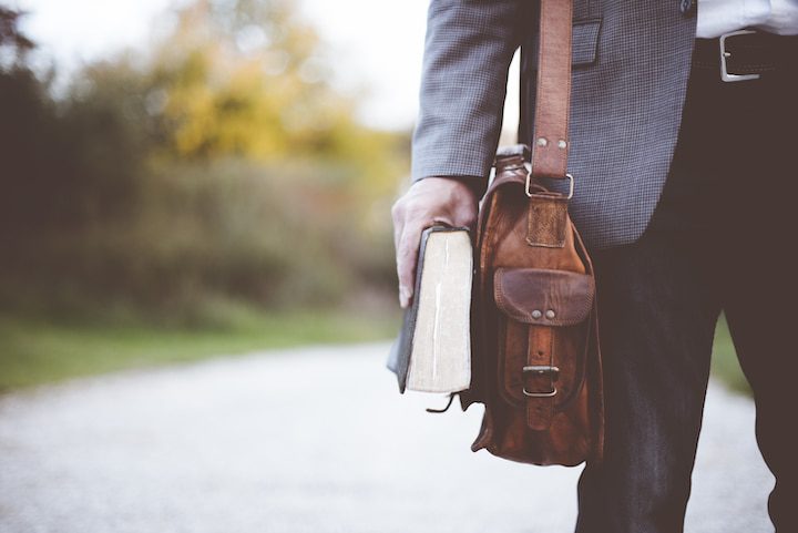 Person holding a book and a brown leather bag, standing on a gravel path outdoors—reflecting on what the Bible says about missions and purposeful journeys.