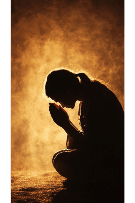 Una persona con silueta se arrodilla bajo una luz cálida y brumosa, con las manos juntas en tranquila contemplación, haciéndose eco del perdón a través de las oraciones que se encuentran en la Biblia.