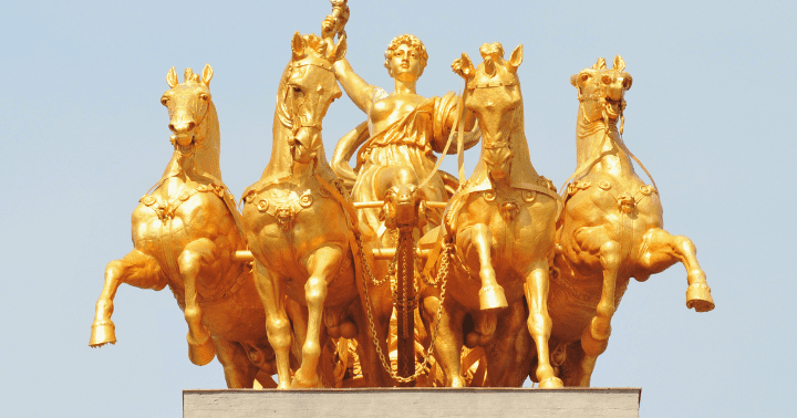 Golden statue of a chariot led by four horses with a figure holding a staff, against a clear sky.