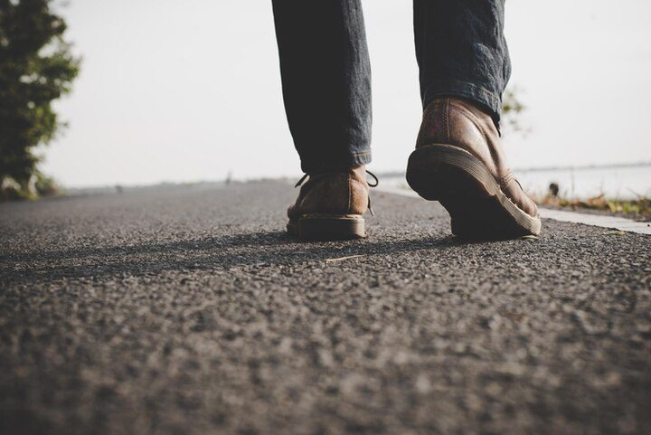 Close-up de uma pessoa caminhando em uma estrada de asfalto, usando sapatos marrons e calças escuras - refletindo a jornada de fé e propósito, ecoando o que a Bíblia diz sobre missões e dando um passo à frente para servir aos outros.