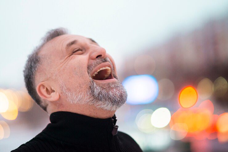 Older man with a gray beard smiling and laughing outdoors, colorful lights blurred in the background, capturing the joy found in laughter in the Bible.