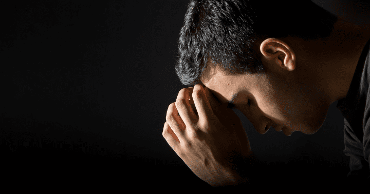 Man with short dark hair, head bowed, and hands clasped in front of his face against a dark background.
