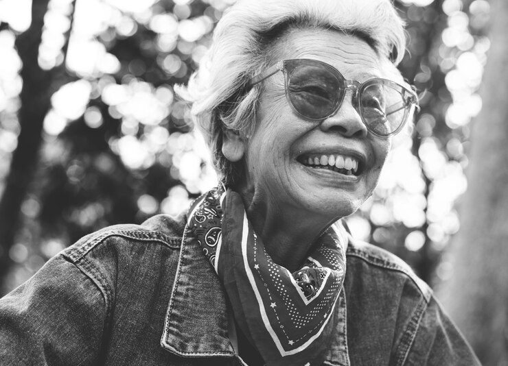 Pessoa idosa sorridente usando óculos escuros, jaqueta jeans e um lenço estampado ao ar livre, irradiando alegria que lembra o riso da Bíblia.